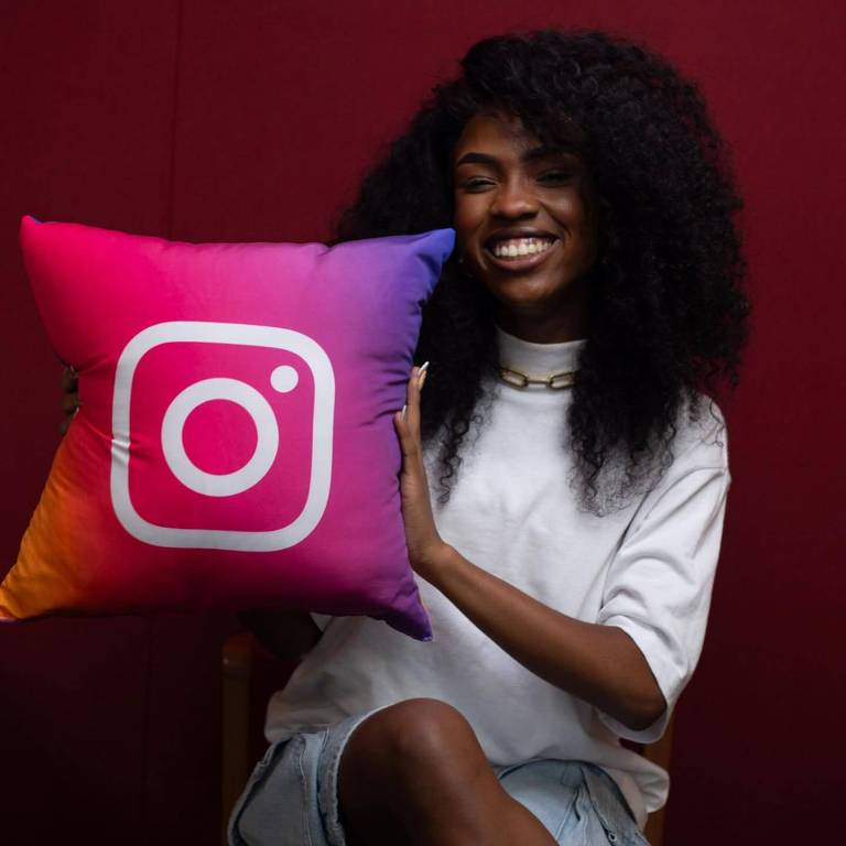 Uma pessoa sorridente está segurando uma almofada com o logotipo do Instagram. A almofada é colorida em um gradiente de rosa e roxo, com o logotipo branco no centro. A pessoa usa uma camiseta branca e um colar dourado, e tem cabelo cacheado e volumoso. O fundo é de uma cor vermelha sólida.