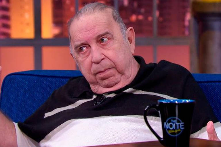 Em foto colorida, homem aparece sentado em um sofá durante uma entrevista a um programa de TV