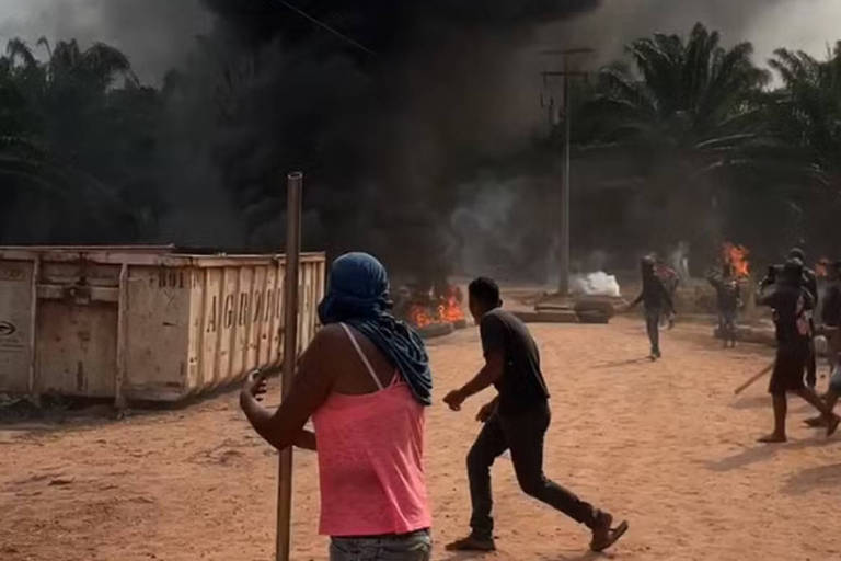 A imagem mostra uma cena de conflito em uma área urbana. No primeiro plano, uma mulher com uma blusa rosa e shorts jeans está segurando um bastão. Ela está descalça e usa um lenço na cabeça. Ao fundo, há fumaça preta densa subindo, indicando um incêndio, e várias pessoas estão se movendo rapidamente, algumas em direção ao fogo. O chão é de terra e há contêineres metálicos visíveis.