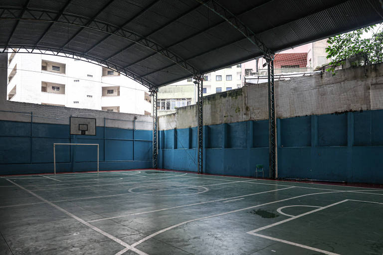 A imagem mostra uma quadra de basquete coberta， com um teto metálico e paredes azuis. No fundo， há um aro de basquete montado na parede. O piso da quadra é verde e apresenta algumas marcas de uso. Ao fundo， são visíveis prédios residenciais e uma área com vegetação.