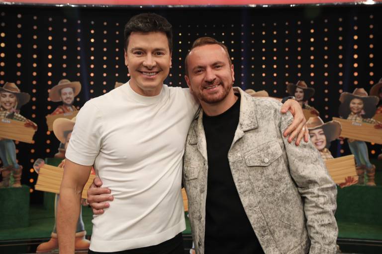 Dois homens estão sorrindo e posando juntos em um estúdio. O primeiro homem, à esquerda, usa uma camiseta branca e está com o braço em volta do segundo homem, que está à direita, vestindo uma jaqueta cinza. Ao fundo, há recortes de figuras de pessoas com chapéus e placas, sugerindo um ambiente de programa de televisão.