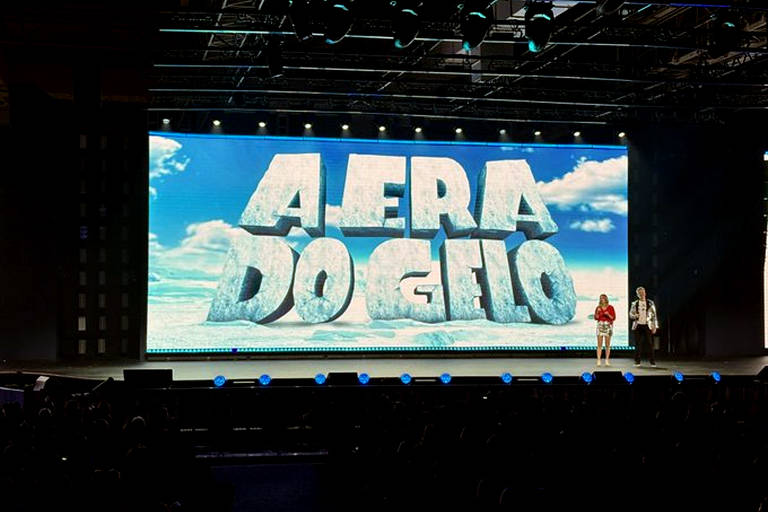 A imagem mostra um palco com uma grande tela ao fundo exibindo a frase 'AERA DO GELO' em letras grandes e tridimensionais, com um fundo de céu azul e nuvens. À frente da tela, duas pessoas estão em pé, uma vestindo um traje vermelho e a outra com um traje prateado. O público é visível na parte inferior da imagem, com uma iluminação suave no palco.