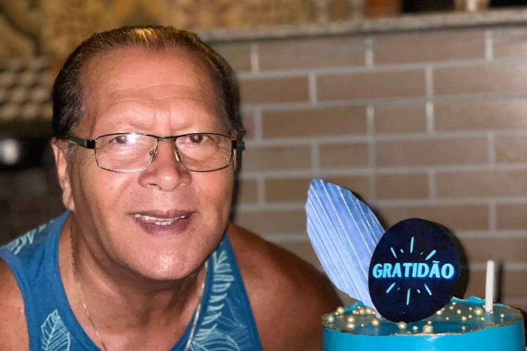 Um homem sorridente está posando ao lado de um bolo de aniversário azul, decorado com detalhes em amarelo e uma vela. O bolo está em uma mesa coberta com uma toalha colorida. Ao lado do bolo, há brigadeiros. O ambiente parece festivo, com uma parede de tijolos ao fundo.