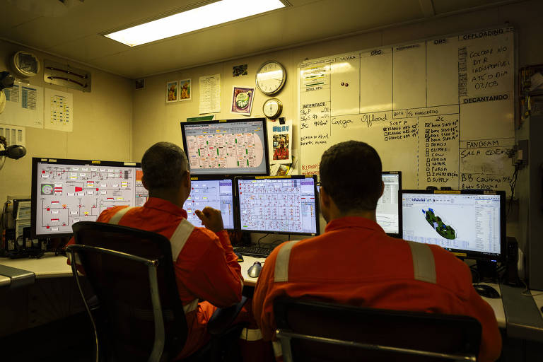 A imagem mostra duas pessoas sentadas em cadeiras de escritório， vestindo uniformes laranja. Elas estão em frente a várias telas de computador que exibem gráficos e dados. O ambiente é uma sala de controle， com paredes cobertas de documentos e um relógio visível. A iluminação é suave， e a atmosfera parece ser de concentração e monitoramento.