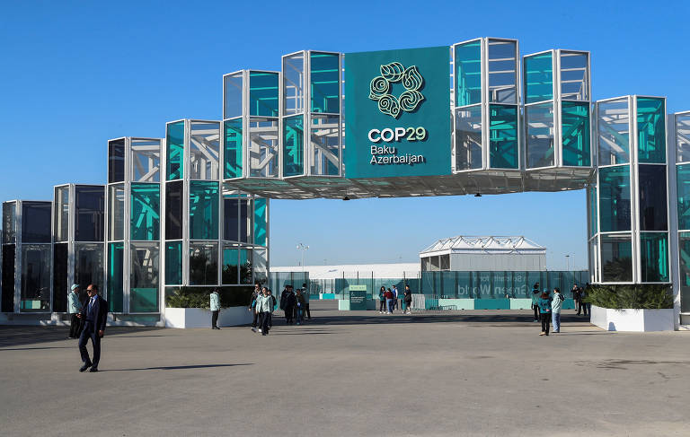 A imagem mostra a entrada do evento COP29, localizada em Baku, Azerbaijão. A estrutura é moderna, composta por painéis de vidro e metal, com o logotipo do evento em destaque. O céu está limpo e azul, e várias pessoas estão caminhando em direção à entrada.