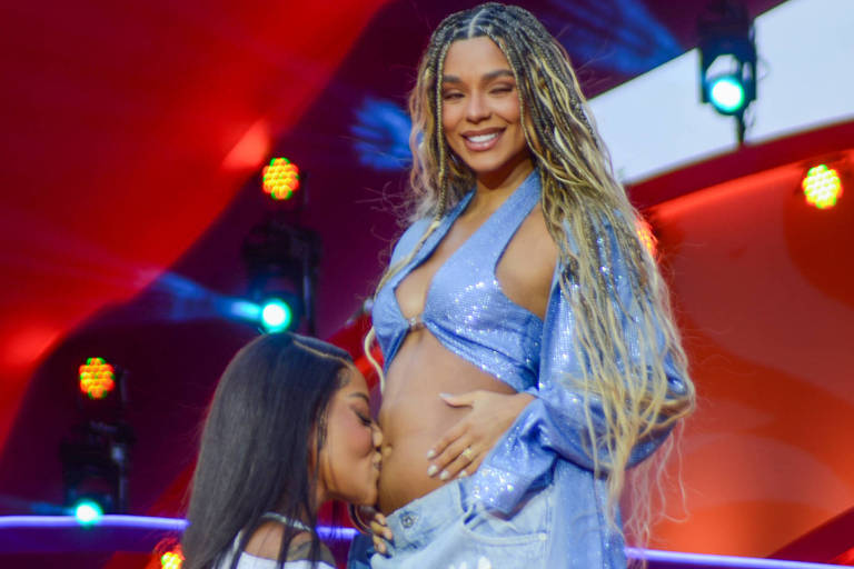 A imagem mostra duas mulheres em um palco. Uma delas, com cabelo longo e trançado, está em pé, usando um top brilhante e calças largas. A outra mulher, de cabelo liso e escuro, está ajoelhada, beijando a barriga da mulher em pé. Ao fundo, há músicos e luzes coloridas, criando um ambiente festivo.