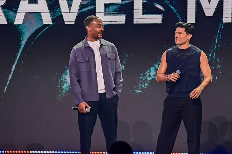 Dois homens estão em um palco durante um evento. O homem à esquerda usa uma camisa cinza e uma camiseta branca, segurando um microfone. O homem à direita está vestido com um colete sem mangas azul escuro e calças pretas, também segurando um microfone.