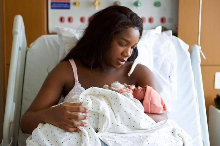 Uma mulher negra sentada em uma cama de hospital, amamentando um recém-nascido. Ela está usando uma blusa de alças e calças de pijama rosa. O bebê está envolto em um cobertor branco com bolinhas e usa um gorro rosa. O fundo mostra equipamentos médicos e uma parede de madeira.