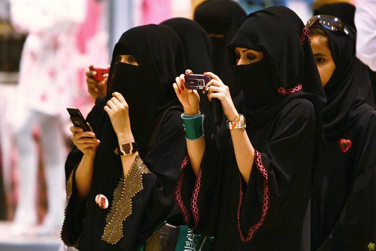 A imagem mostra um grupo de mulheres vestindo abayas e niqabs, segurando celulares e tirando fotos. Elas estão em um ambiente com um fundo desfocado. As mulheres estão em pé, algumas com acessórios visíveis, como pulseiras e relógios.