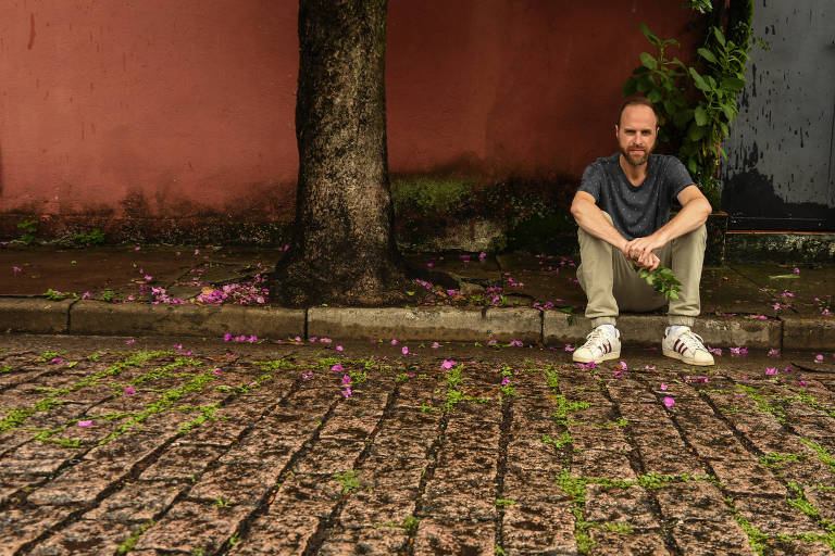 Um homem está sentado no chão de um espaço urbano, próximo a uma árvore. O fundo é uma parede de cor rosa e o chão é coberto por pedras. Há flores pequenas e roxas espalhadas ao redor do homem, que usa uma camiseta escura e calças claras. Ele parece pensativo.