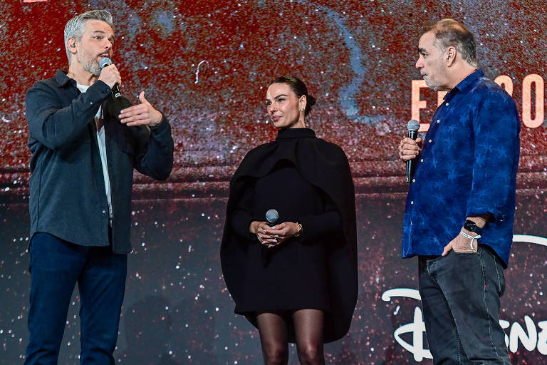 Três pessoas estão em um palco durante um evento. À esquerda, um homem com cabelo grisalho e camisa escura fala ao microfone. No centro, uma mulher com cabelo preso e vestido preto escuta atentamente, segurando um microfone. À direita, um homem com cabelo curto e camisa azul com estampas observa. Ao fundo, há uma tela com o logotipo da Disney+.