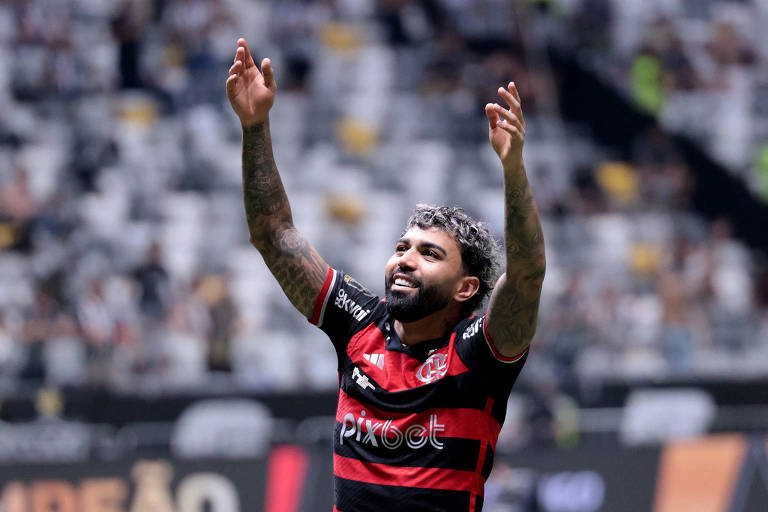 Um jogador de futebol está comemorando com os braços levantados. Ele usa uma camisa listrada em vermelho e preto, com o logo do clube e patrocinadores visíveis. O fundo mostra um estádio com torcedores, mas as arquibancadas estão desfocadas.
