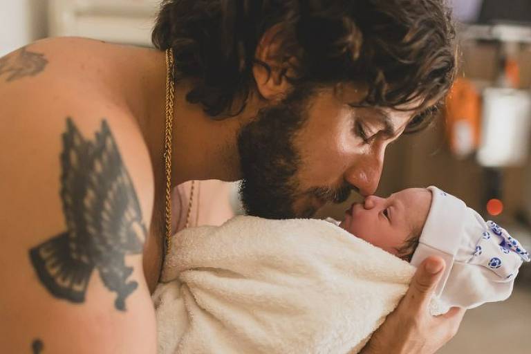 A imagem mostra um homem com cabelo longo e barba, segurando um bebê envolto em uma toalha branca. O homem está se inclinando para frente, tocando o nariz do bebê com o seu, em um gesto carinhoso. Ele está sem camisa e possui várias tatuagens visíveis em seu braço. O ambiente parece ser um quarto, com alguns objetos ao fundo.