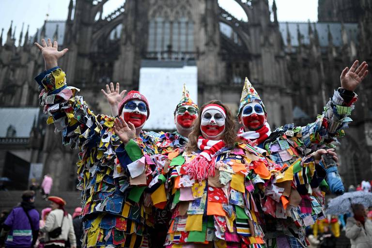 Três palhaços estão posando para a foto em frente a uma catedral. Eles estão vestidos com trajes coloridos feitos de retalhos de tecido e têm rostos pintados com maquiagem de palhaço. Todos estão levantando as mãos e sorrindo, enquanto o fundo apresenta a arquitetura gótica da catedral.