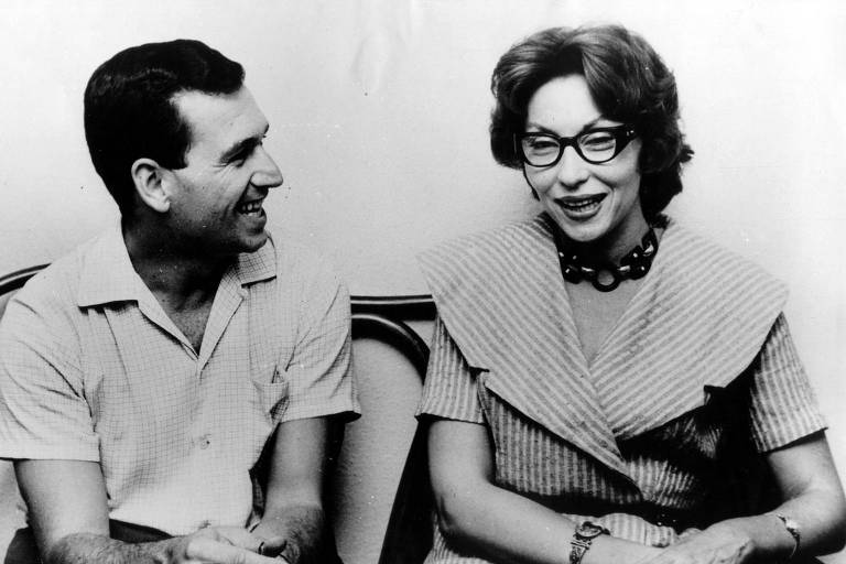 A imagem em preto e branco mostra um homem e uma mulher sentados lado a lado. O homem, à esquerda, está sorrindo e vestindo uma camisa clara. A mulher, à direita, usa óculos e uma blusa com padrão listrado, além de um colar. Ambos parecem estar em uma conversa descontraída.
