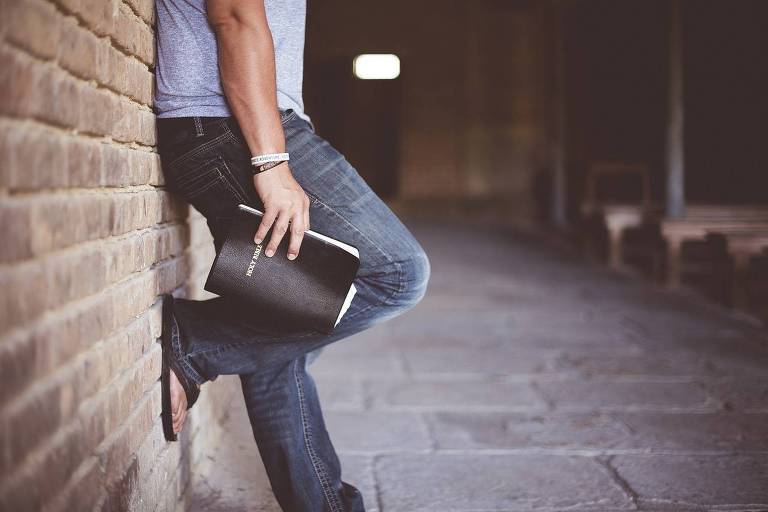 A imagem mostra a parte inferior de uma pessoa encostada em uma parede de tijolos. A pessoa está usando uma camiseta azul e jeans escuros， segurando um caderno preto. O ambiente parece ser um corredor com piso de pedras e uma iluminação suave ao fundo.