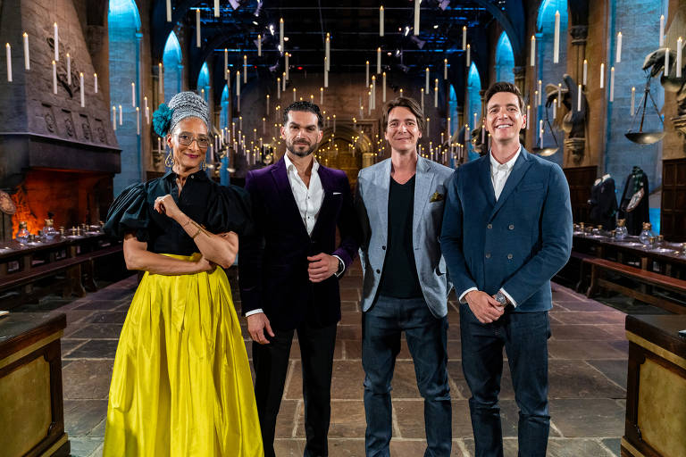 Quatro pessoas posam em um ambiente que parece ser um set de filmagem de um programa de culinária. À esquerda, uma mulher com cabelo preso em um coque e vestido preto com uma saia amarela. Ao lado dela, um homem com terno roxo e camisa branca. O terceiro é um homem com um blazer cinza e camiseta preta. Por último, um homem com um blazer azul claro e camisa branca. O fundo apresenta um ambiente com mesas longas e velas penduradas.