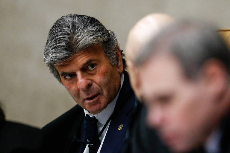 A imagem mostra um homem com cabelo grisalho e bem penteado， vestindo um terno escuro e uma gravata. Ele está olhando para o lado， com uma expressão séria， enquanto participa de uma reunião. Ao fundo， há outras pessoas， mas seus rostos não estão claramente visíveis.
