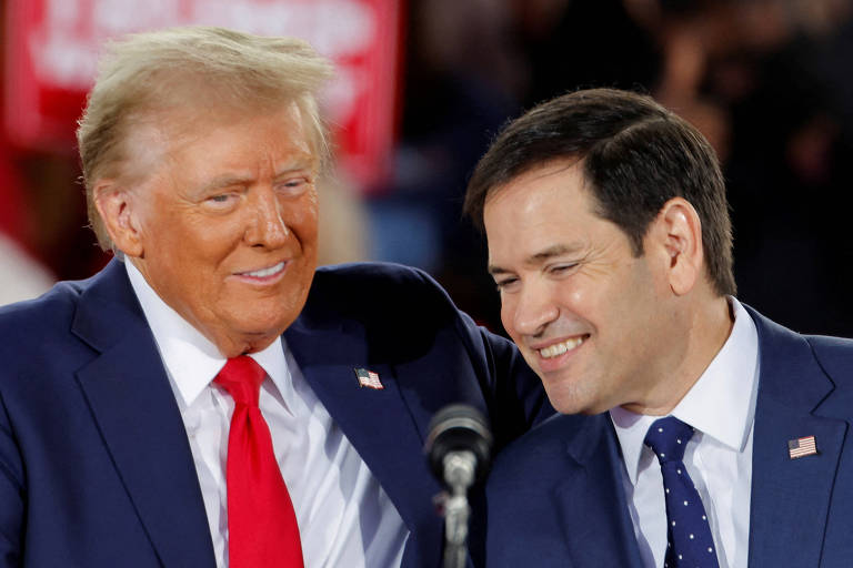 A imagem mostra duas pessoas em um evento político. A pessoa à esquerda tem cabelo loiro e usa um terno escuro com uma gravata vermelha. A pessoa à direita tem cabelo escuro e usa um terno escuro com uma gravata azul com bolinhas brancas. Ambos estão sorrindo e parecem estar se divertindo. Ao fundo， há um sinal vermelho que não é legível