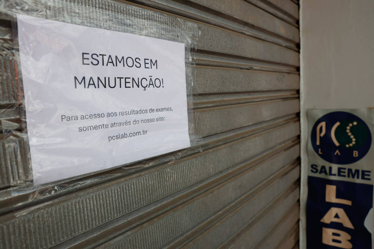 Uma placa branca colada em uma porta de metal fechada, informando que o local está em manutenção. O texto diz: 'ESTAMOS EM MANUTENÇÃO. Para mais informações, entre em contato pelo telefone: (XX) XXXX-XXXX.' Ao lado, há um logotipo que diz 'SALEME LAB'.