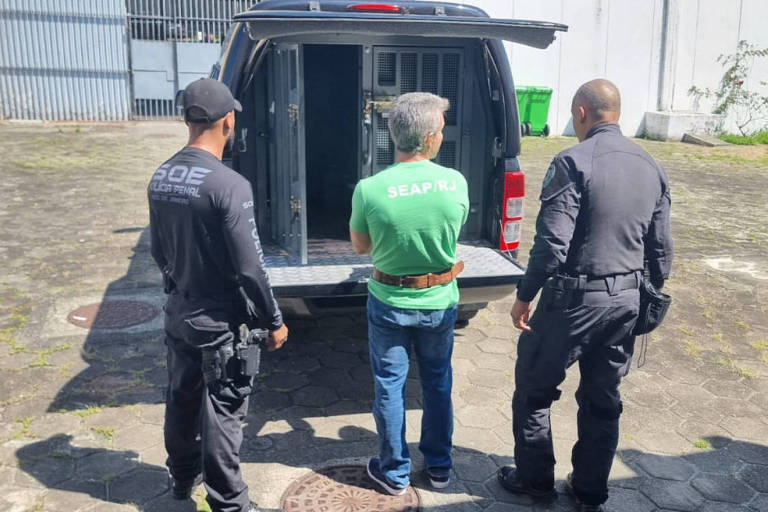 A imagem mostra três homens vistos de costas. Dois deles estão vestidos com uniformes de segurança， um em preto e outro em azul escuro. O terceiro homem， que está no meio， usa uma camiseta verde e está com as mãos amarradas. Eles estão próximos a uma van de polícia， que está com a porta traseira aberta. O ambiente parece ser um espaço externo， com um piso de concreto e algumas estruturas ao fundo.