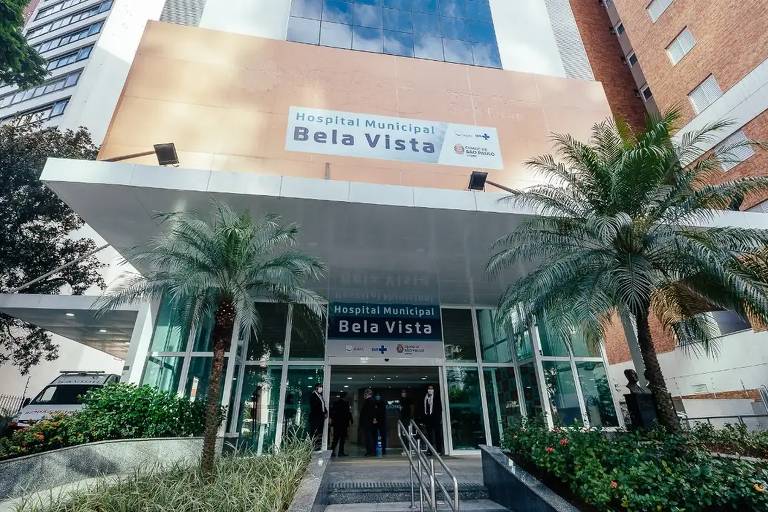 A imagem mostra a entrada do Hospital Bela Vista， com uma fachada moderna. Há um grande letreiro com o nome 039;Hospital Bela Vista039; na parte superior. A entrada é ampla， com escadas e rampas， cercada por plantas e palmeiras. O céu está claro ao fundo.