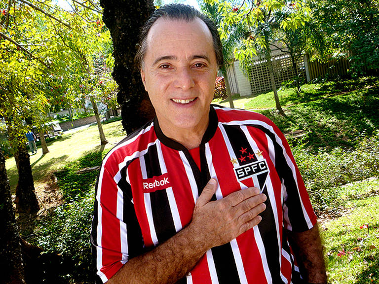 Um homem sorridente está posando ao ar livre, vestindo uma camisa do São Paulo FC, que é predominantemente vermelha e preta com listras verticais. Ele está com a mão sobre o coração, em um gesto de carinho ou respeito. Ao fundo, há árvores e um gramado verde, sugerindo um ambiente natural e tranquilo