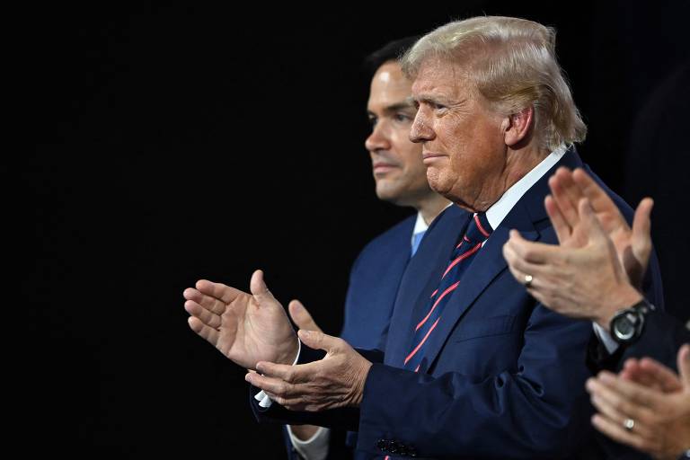 Foto mostra Donald Trump, um homem branco, com linhas de expressões de idade, cabelos grisalhos, louros e com sinais de calvície batendo palmas. ele veste um terno azul-escuro com gravata listrada vermelha e azul e uma camisa branca. O fundo da imagem está escuro e ao redor dele outras pessoas brancas batem palmas.