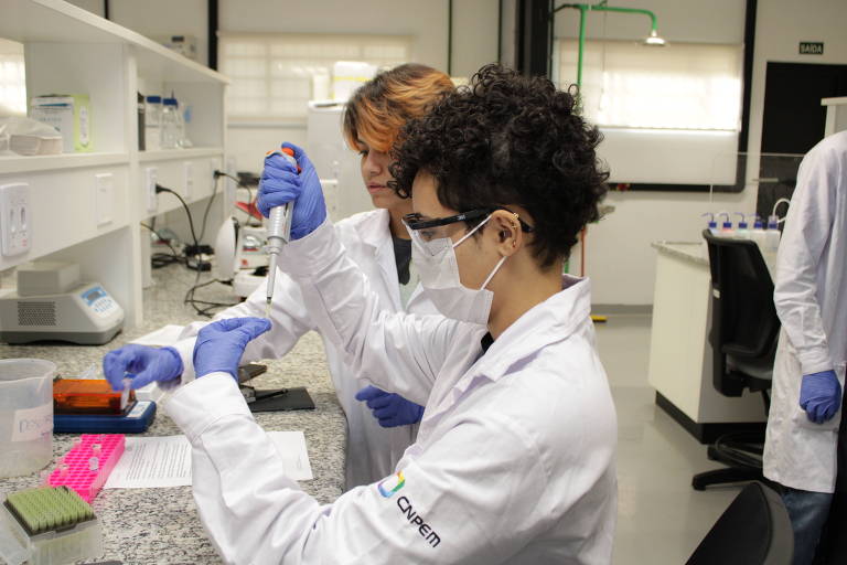 A imagem mostra duas pessoas em um laboratório. Uma delas， com cabelo cacheado e óculos， está usando um pipetador， enquanto a outra， com cabelo liso， observa e auxilia. Ambas estão vestindo jalecos brancos e luvas azuis. O ambiente é bem iluminado， com bancadas e equipamentos de laboratório ao fundo.