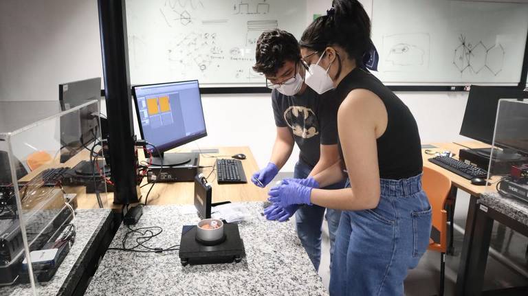 Dois estudantes estão em um laboratório, usando máscaras e luvas. Um deles está segurando um objeto enquanto o outro observa. Ao fundo, há computadores e uma lousa com anotações. A mesa é coberta por uma superfície de granito.