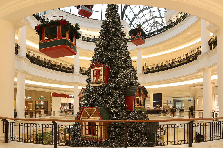 A imagem mostra uma grande árvore de Natal decorada com luzes e enfeites, localizada no centro de um shopping. A árvore possui casas decorativas em sua base e está cercada por um corrimão. Acima da árvore, há cabines de teleférico decoradas com folhagens e laços vermelhos. O ambiente é iluminado e apresenta lojas ao fundo, com uma arquitetura moderna e elegante.