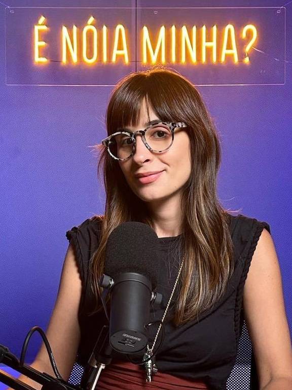 A imagem mostra uma mulher com cabelo longo e liso, usando óculos com armação clara. Ela está sentada em frente a um microfone em um estúdio, com um fundo azul. Acima dela, há uma placa iluminada com a frase 'É nóia minha?'. A mulher está vestindo uma blusa preta e parece estar sorrindo levemente.