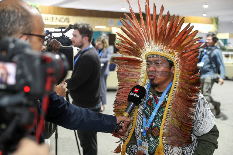 A imagem mostra um líder indígena usando um elaborado cocar de penas, sendo entrevistado por um repórter. O ambiente parece ser um evento ou conferência, com outras pessoas ao fundo, algumas com câmeras. O líder está vestido com roupas tradicionais e segura um microfone enquanto fala.