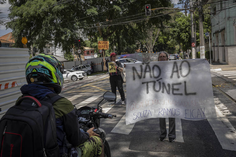 A imagem mostra um protesto na rua， onde uma pessoa está segurando um cartaz que diz 039;NÃO AO TÚNEL039;. Um motociclista está parado em frente ao protesto， observando a cena. Ao fundo， há árvores e veículos estacionados.