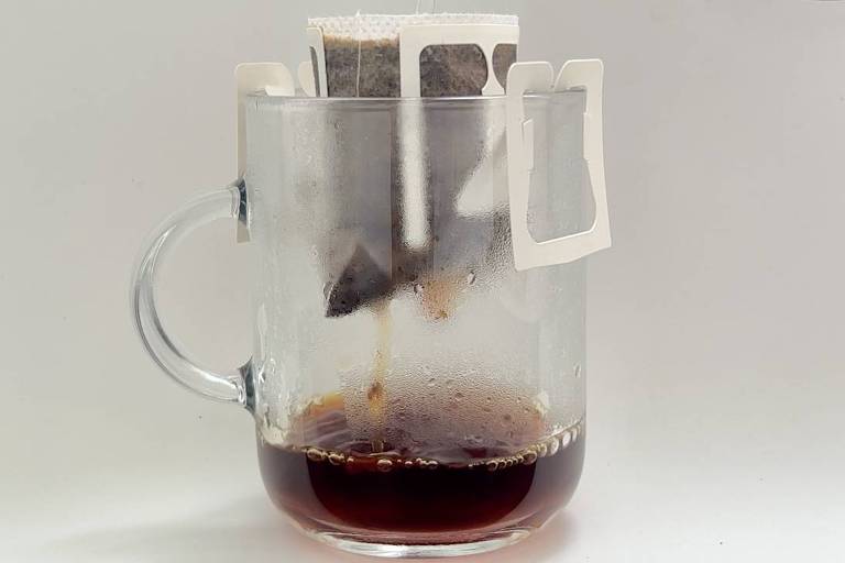 A imagem mostra um copo de vidro transparente com café sendo preparado. Um filtro de café está posicionado na borda do copo， com água quente sendo despejada sobre ele. O café já preparado está visível na parte inferior do copo， que apresenta uma coloração escura. O fundo da imagem é branco， e há uma superfície preta na parte inferior.