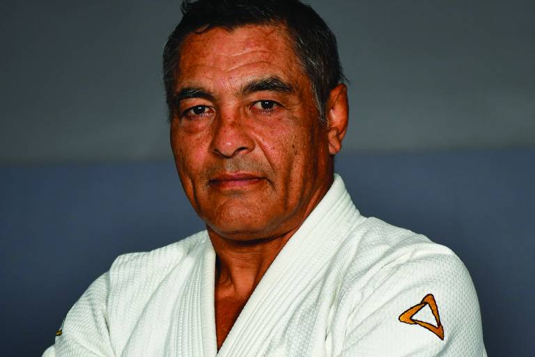 Um homem idoso está posando com os braços cruzados, vestindo um quimono branco de Jiu-Jitsu. Ele tem cabelo escuro e levemente grisalho, e sua expressão é séria. O quimono possui um emblema na parte esquerda do peito e uma faixa laranja na manga direita. O fundo é de cor cinza.