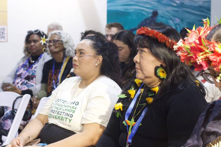 Duas mulheres indígenas estão sentadas uma ao lado da outra. Uma delas usa camisa branca e pintura facial, a outra usa preso e uma tiara de penas na cabeça. 