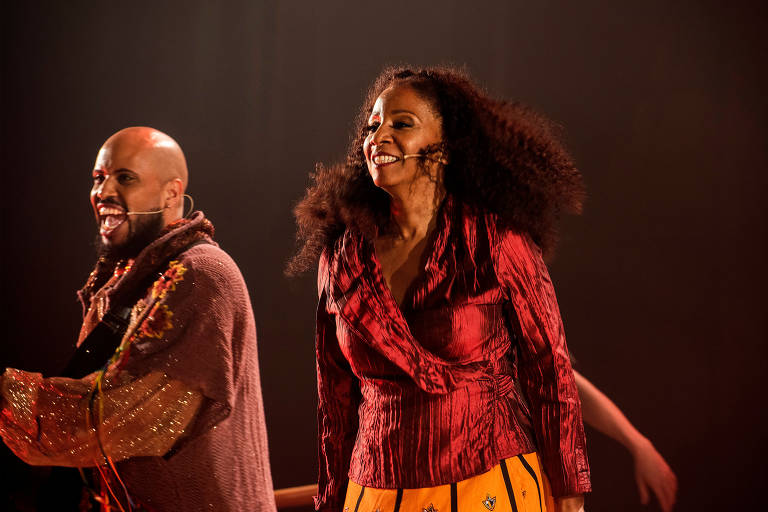 A imagem mostra dois artistas em uma apresentação no palco. À esquerda, um homem com cabelo raspado e barba, vestindo uma roupa de tom marrom e detalhes brilhantes, sorri enquanto segura um instrumento. À direita, uma mulher com cabelo cacheado e volumoso, usando uma blusa vermelha e uma saia amarela com estampas, também sorri. O fundo é escuro, com iluminação suave que destaca os artistas.