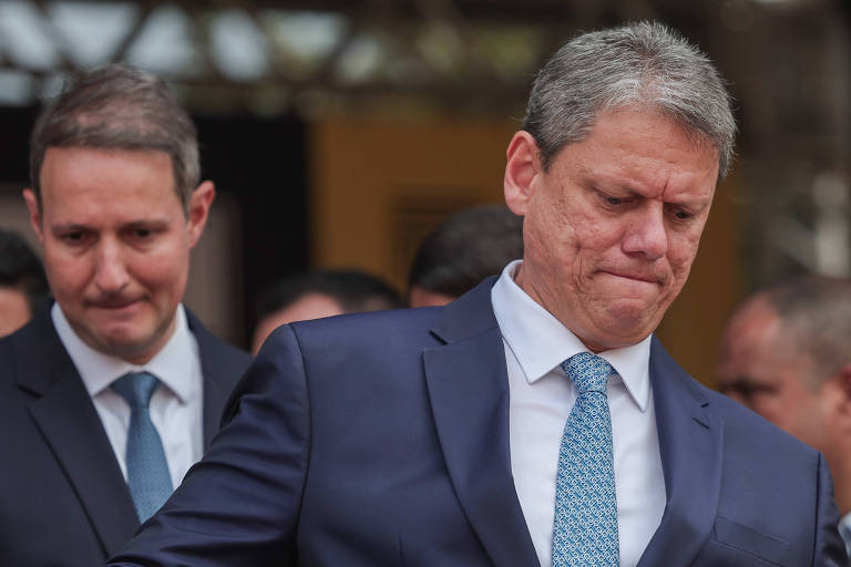 A imagem mostra dois homens em trajes formais. O homem à frente é Tarcísio, com cabelo grisalho, está usando um terno escuro e uma gravata clara, com uma expressão de preocupação. O homem ao fundo, Derrite, com cabelo mais escuro, também está vestido formalmente e observa o homem à frente. O ambiente parece ser ao ar livre, com pessoas ao fundo.