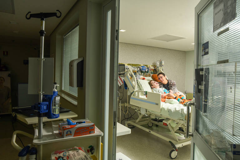 A imagem mostra uma criança em um leito hospitalar, com uma mulher ao seu lado. A criança parece estar deitada, com um olhar tranquilo, enquanto a mulher sorri para ela. O ambiente é um quarto de hospital, com equipamentos médicos visíveis ao fundo, como monitores e um suporte para soro. Há uma mesa com alguns itens, incluindo uma caixa e um recipiente. A luz natural entra pela janela, iluminando o espaço.