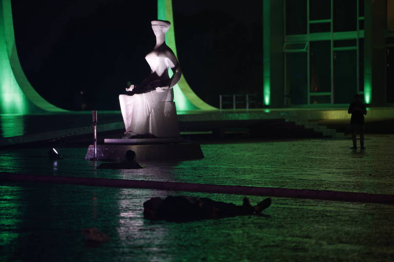 A imagem mostra uma escultura em um espaço aberto, iluminada por luzes verdes. No primeiro plano, há um corpo no chão, enquanto ao fundo, uma pessoa está em pé, observando a escultura. O ambiente é noturno, com reflexos no chão molhado.