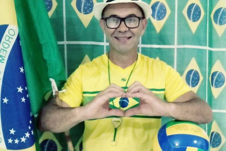 Um homem está sentado em uma cadeira, vestindo uma camiseta amarela e calças verdes, representando as cores da bandeira do Brasil. Ele faz um gesto de coração com as mãos, simbolizando amor pelo país. Ao seu lado, há uma bola de vôlei e uma bandeira do Brasil ao fundo, que é decorada com o padrão da bandeira nacional.