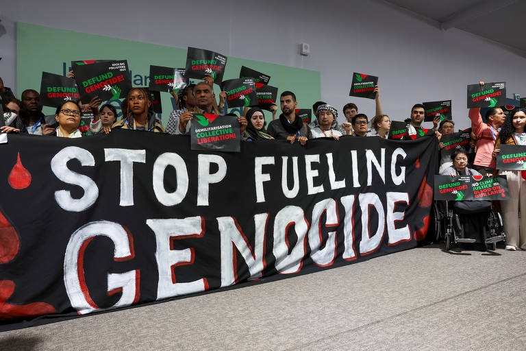 Um grupo de pessoas exibe cartazes com os dizeres "demilitarisation collective liberation", posicionados atrás de uma faixa escrito "stop fueling genocide".