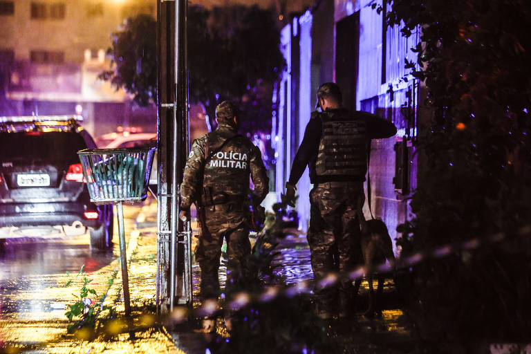 A imagem mostra dois policiais militares caminhando em uma rua durante uma tempestade. Eles estão vestidos com uniformes camuflados e coletes à prova de balas. Ao fundo, há um carro estacionado e a iluminação da rua é refletida em poças de água no chão. A cena é noturna e a atmosfera é de chuva intensa.