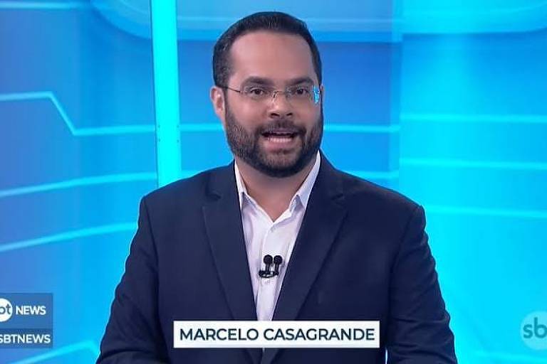 Um apresentador de telejornal está em um estúdio com fundo azul. Ele usa um terno escuro e uma camisa branca. O apresentador tem barba e óculos, e está sorrindo enquanto fala. Na parte inferior da imagem, está escrito 'MARCelo CASAGRANDE' em uma faixa branca. No canto inferior esquerdo, há o logotipo do SBT News.