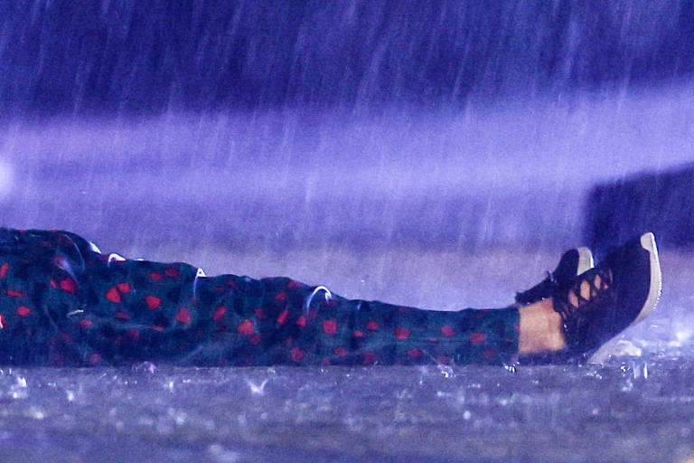 Na chuva à noite, estendido no chão, o corpo de um homem, que aparece das pernas para baixo, vestido com uma calça verde com bolinhas vermelhas e tênis pretos