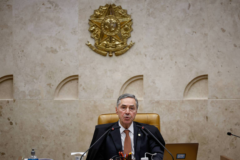 Um homem sentado em uma cadeira de couro em um ambiente oficial, com uma parede de fundo em tom claro e um brasão dourado acima dele. O homem está usando um terno escuro e uma gravata, e parece estar falando ou apresentando algo. Na mesa à sua frente, há alguns objetos, incluindo uma garrafa de água e um copo.