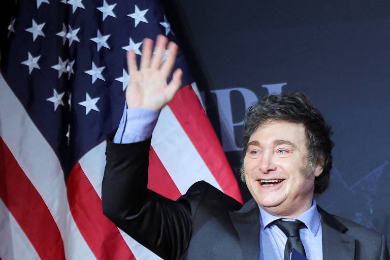 Um homem com cabelo escuro e ondulado， vestindo um terno preto e uma camisa azul， está acenando com a mão direita. Ao fundo， há bandeiras dos Estados Unidos. O homem parece estar sorrindo e se dirigindo a uma audiência.
