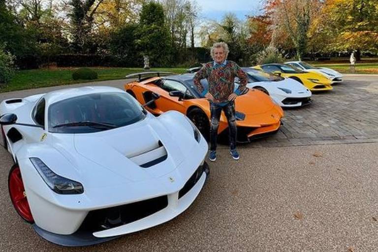 Em foto colorida, homem posa com sua coleção de cinco carros esportivos no quintal de sua casa