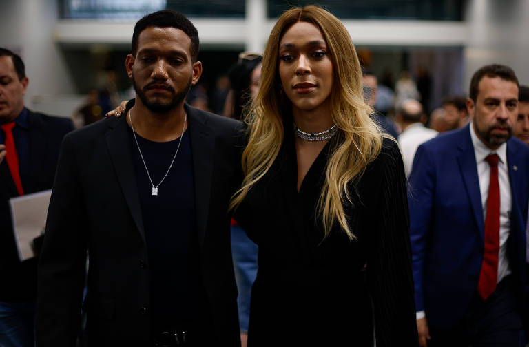 A imagem mostra duas pessoas caminhando juntas em um ambiente interno. A pessoa à esquerda é um homem com cabelo curto e barba, vestindo uma camiseta preta e um blazer. Ele está segurando o braço da mulher à direita, que tem cabelo longo e liso, usando um vestido preto e colar. Ao fundo, há outras pessoas e uma iluminação suave.